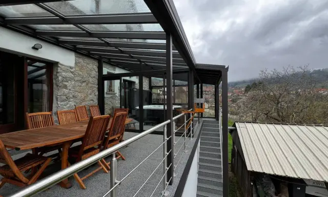 Veranda sur terrasse surélevée avec jacuzzi et table en bois. Vue sur un village et des collines brumeuses.