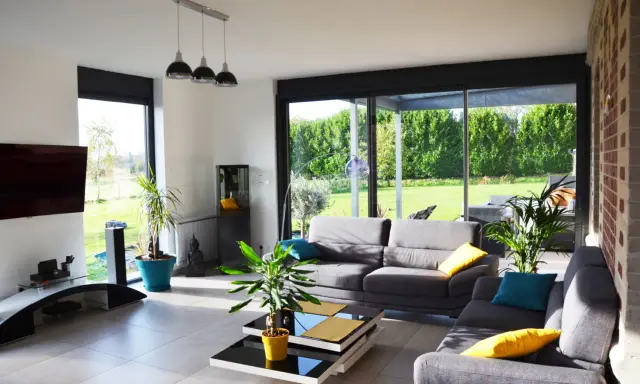 Salon lumineux avec canapés gris, table basse moderne et grandes baies vitrées donnant sur un jardin verdoyant. Une annexe maison parfaite pour se détendre.