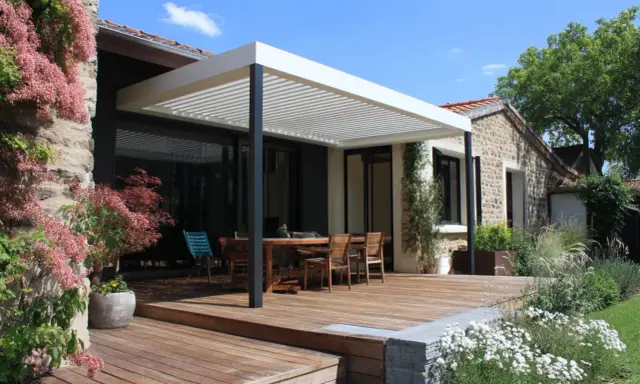 Terrasse en bois sous un toit pergola naturel, avec table et chaises, entourée de végétation luxuriante et d'une maison en pierre.