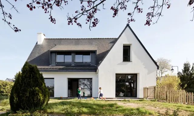 Une extension maison néo-bretonne moderne avec de grandes baies vitrées. Deux enfants jouent dans le jardin verdoyant devant la maison.