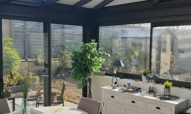 Intérieur lumineux d'une véranda triple vitrage, avec vue sur un jardin verdoyant et une commode blanche décorée.