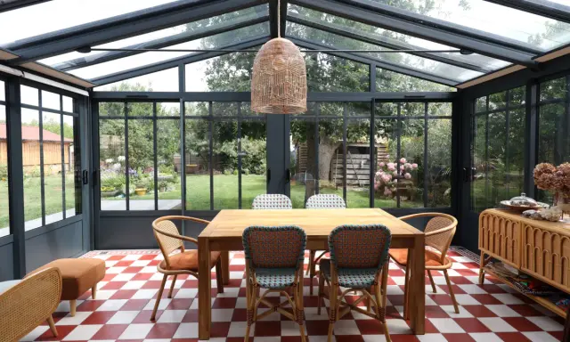 Salle à manger lumineuse dans une véranda avec toiture zinc. Table en bois, chaises colorées, sol à damier rouge et blanc, vue sur jardin verdoyant.