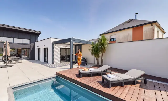 Une femme en robe colorée se tient près d'une pergola moderne, profitant de l'étanchéité pergola mur. Piscine et chaises longues complètent ce paysage de détente.