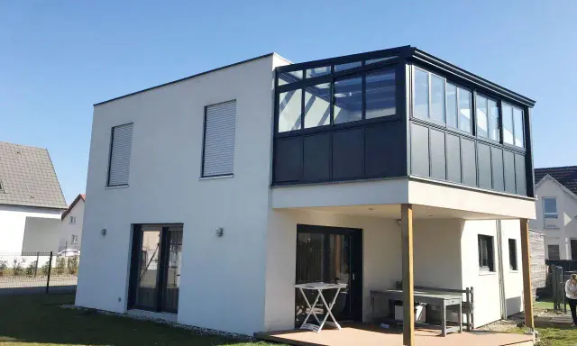 Maison moderne avec extension étage sur piloti. Une véranda vitrée surplombe une terrasse en bois.