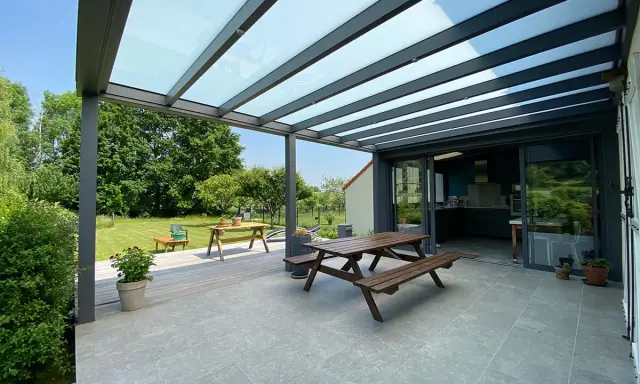 Terrasse avec table en bois sous une pergola bioclimatique. La hauteur de la structure permet de profiter du jardin verdoyant.