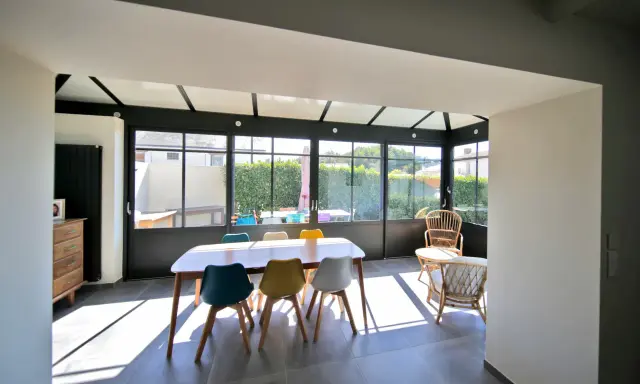 Salle à manger lumineuse dans une véranda chauffée, avec une table en bois et des chaises colorées. Un espace de vie agréable.