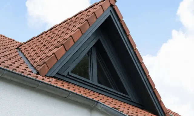 Un puit de lumière triangulaire s'intègre dans un toit en tuiles rouges, sous un ciel bleu et nuageux.