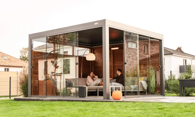 Un couple se détend dans une pergola moderne avec parois vitrées. La fixation pergola est discrète, laissant place à un espace cosy avec chauffage d'appoint.