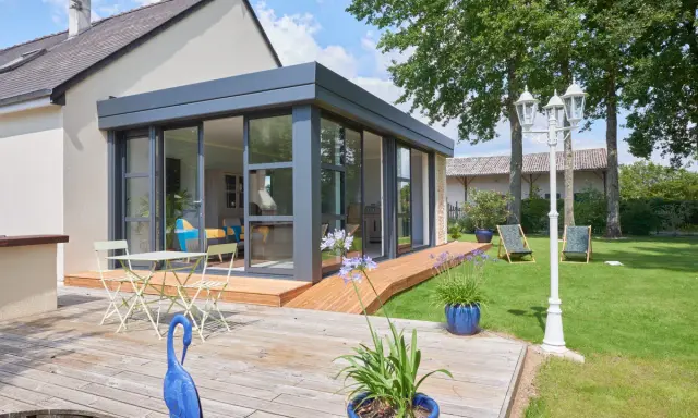 Extension toit plat sur maison ancienne, avec terrasse en bois, mobilier de jardin et vue sur un jardin verdoyant.