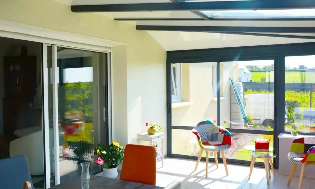 Intérieur lumineux d'une véranda avec un grand vitrage offrant une vue sur un jardin verdoyant. Des chaises colorées invitent à la détente.