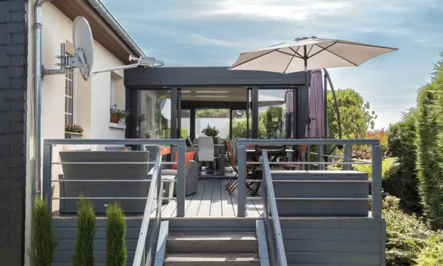 Terrasse avec mobilier de jardin et une véranda bois et alu. Parasol ouvert, escalier menant à la terrasse.