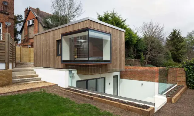 Une extension de maison moderne en bois avec une grande fenêtre en saillie. L'agrandissement maison s'intègre à un jardin en pente avec des murs de briques et des garde-corps en verre.