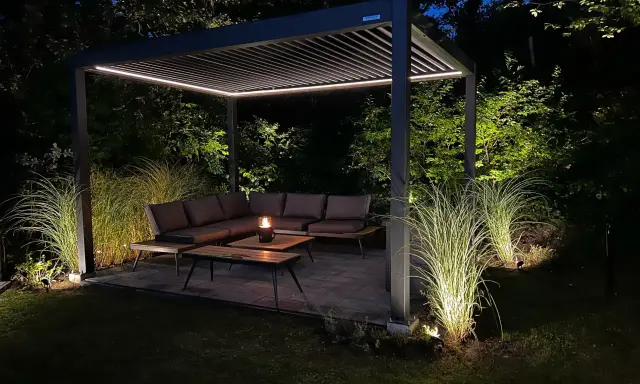 Une **pergola au milieu du jardin** illuminée la nuit, avec un salon de jardin confortable et des herbes hautes éclairées.