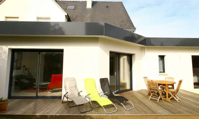 Extension de toit plat en bac acier, avec terrasse en bois, chaises longues et table de jardin.