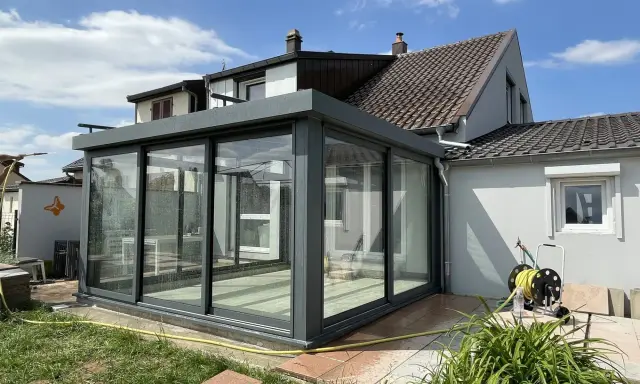 Une véranda moderne, conçue par un architecte, prolonge une maison. Elle est en verre et métal gris, avec un toit plat. Le jardin est verdoyant.