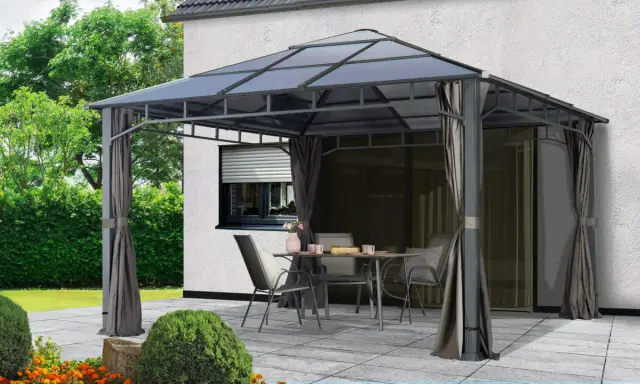 Une tonnelle def. avec table et chaises sur une terrasse pavée, devant une maison blanche et un jardin verdoyant.
