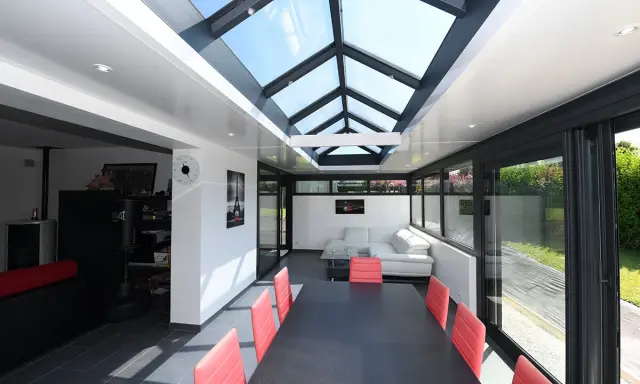 Une extension veranda salle à manger moderne avec un toit vitré, une table noire et des chaises rouges, un canapé blanc et une vue sur le jardin.