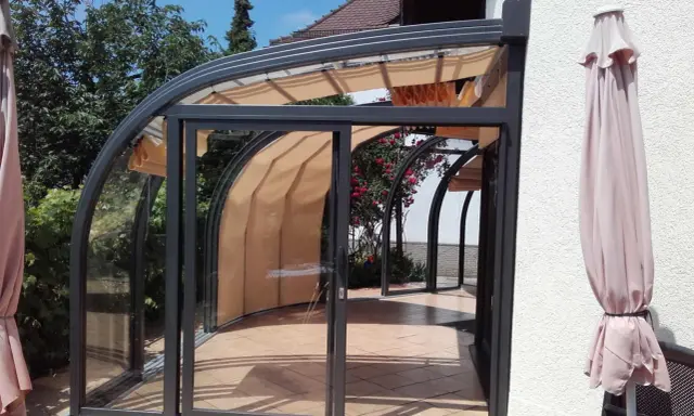 Une élégante veranda demi ronde en verre et métal, avec des stores rétractables, s'ouvre sur un jardin fleuri.