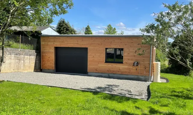 Extension garage bois accolé maison, avec porte noire, fenêtre et jardin verdoyant.