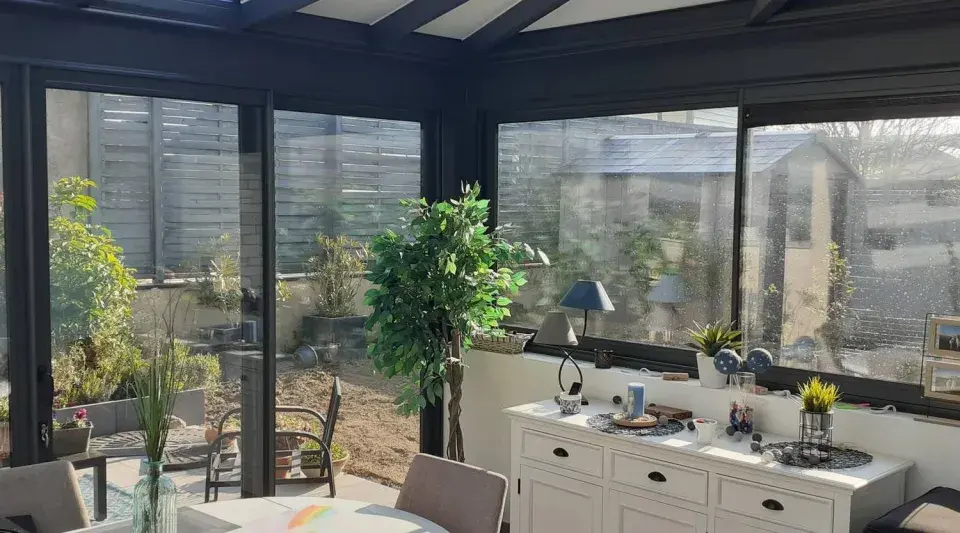 Intérieur lumineux d'une véranda triple vitrage, avec vue sur un jardin verdoyant et une commode blanche décorée.