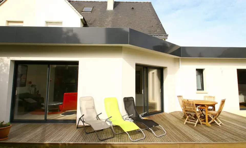 Extension de toit plat en bac acier, avec terrasse en bois, chaises longues et table de jardin.