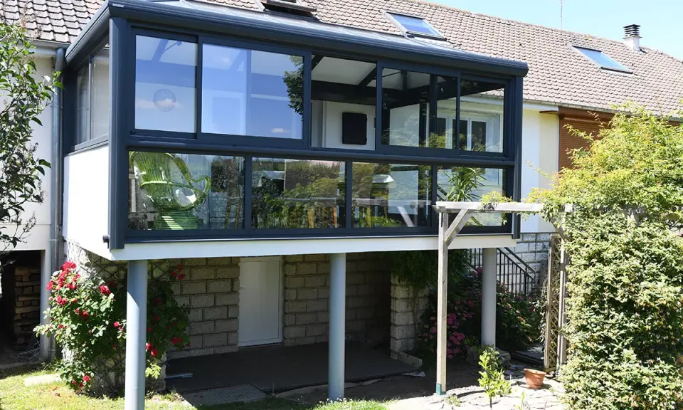 Moderne veranda adossée au mur, avec une chaise verte visible à travers les grandes baies vitrées. Le soleil brille sur le jardin verdoyant.