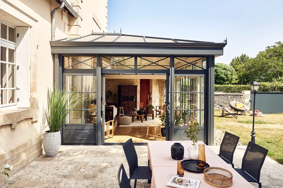 Une véranda moderne s'ouvre sur un jardin verdoyant. Une table dressée invite à profiter de l'extérieur, peut-être pour éviter la taxe terrasse.