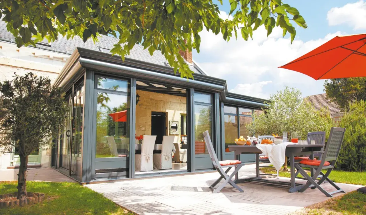 Une véranda moderne avec un surbot béton, une table et des chaises sur une terrasse en bois, sous un parasol orange vif.