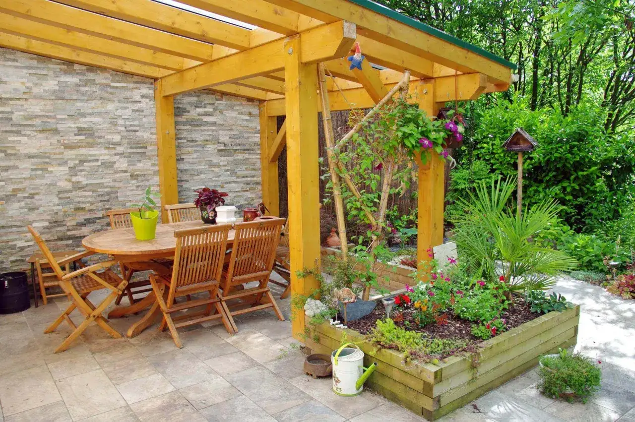 Table et chaises en bois sous une pergola &agrave; fixation solide, entour&eacute;es d'un jardin fleuri et d'un mur en pierre.