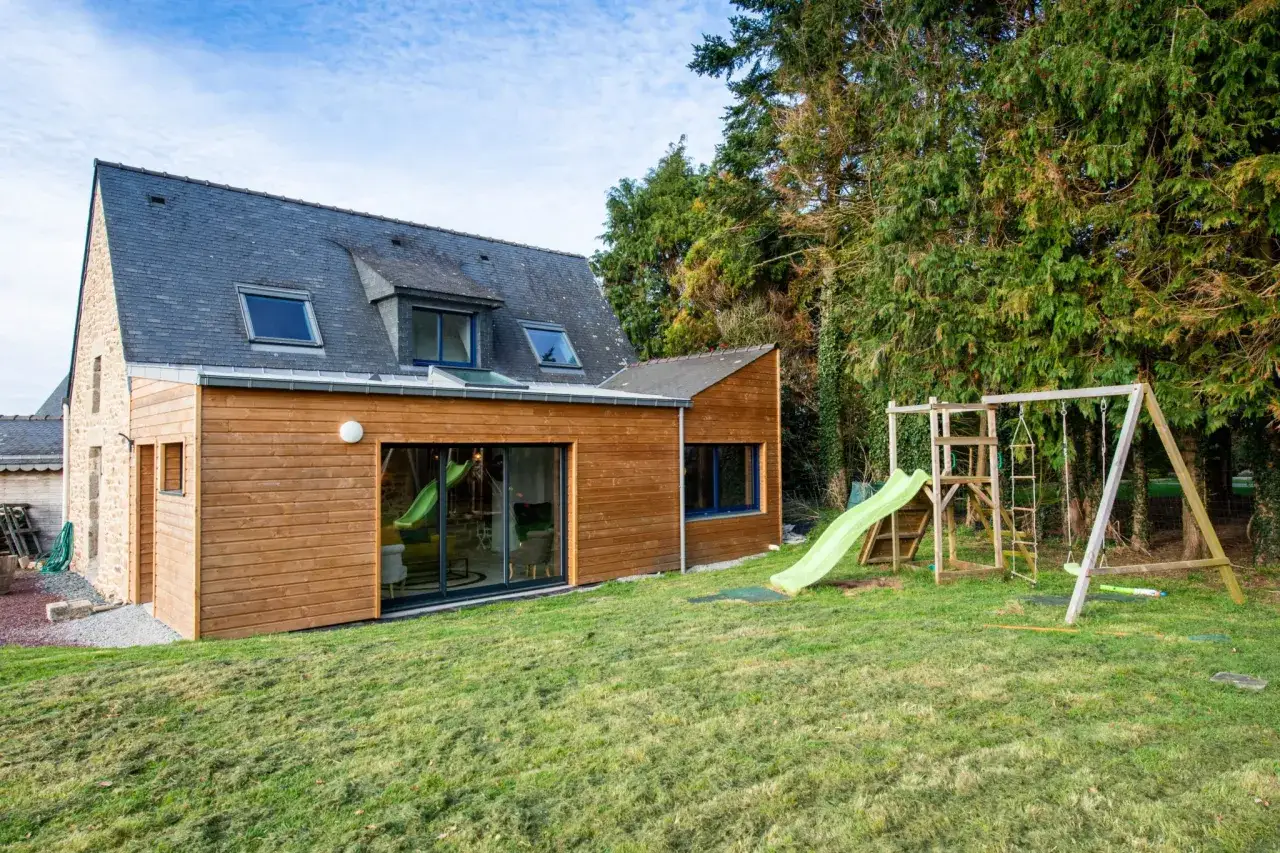 Agrandissement d'un chalet en bois avec une extension moderne, un jardin verdoyant et une aire de jeux pour enfants.