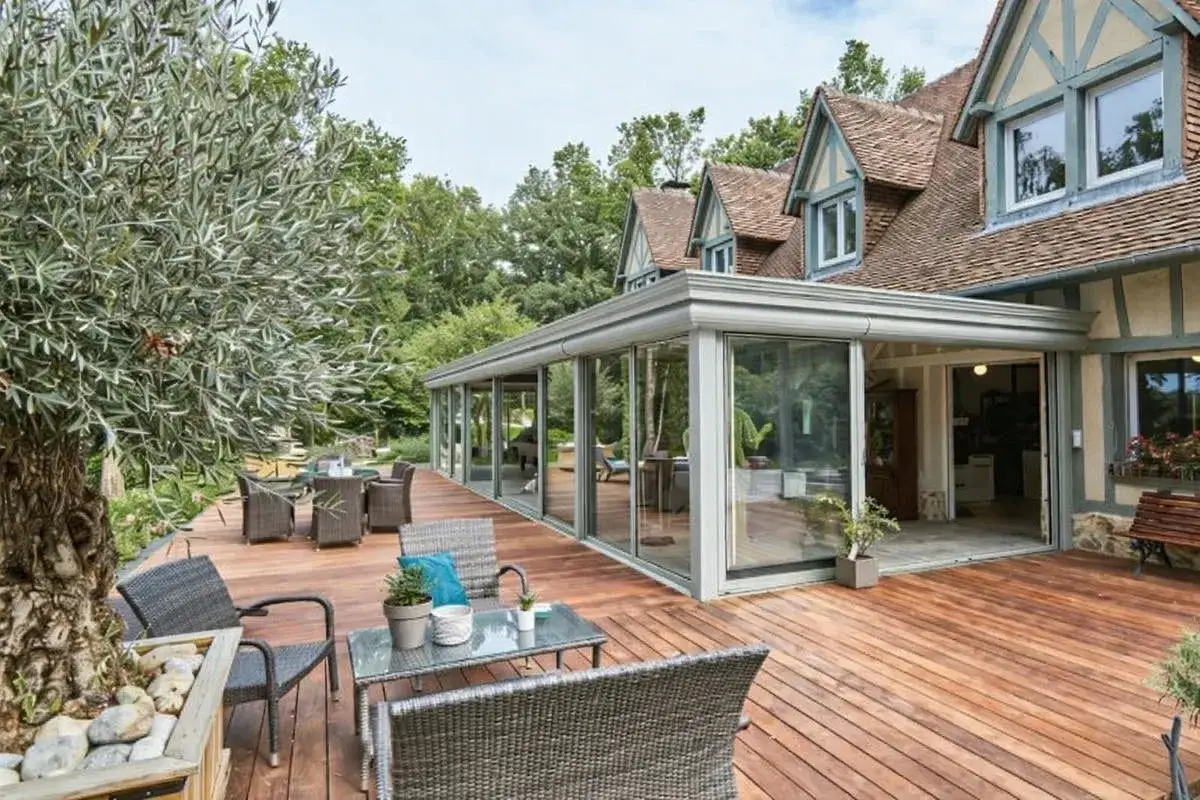 Une extension habitation moderne en verre s'intègre à une maison traditionnelle. Terrasse en bois avec mobilier d'extérieur.