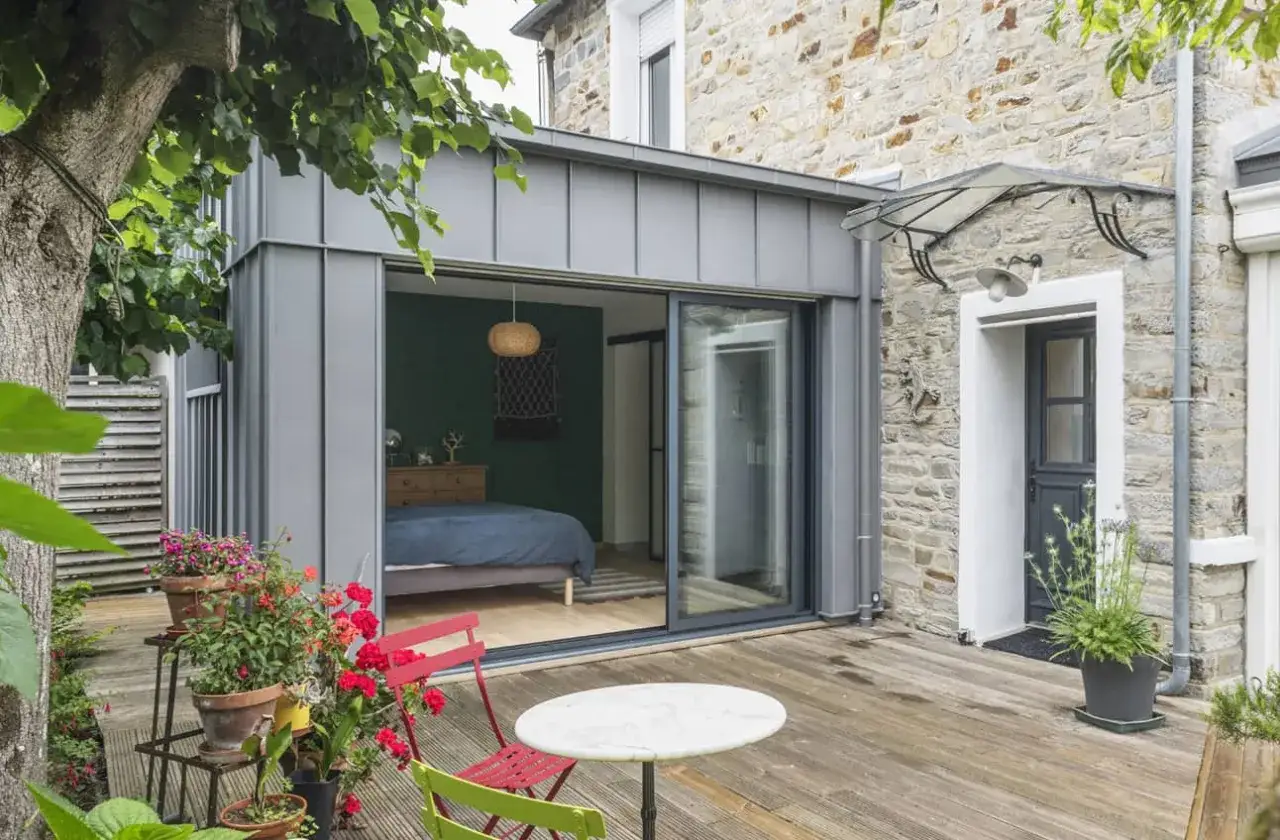 Une extension maison style atelier avec une grande baie vitrée s'ouvrant sur une terrasse en bois. Un lit bleu est visible à l'intérieur.