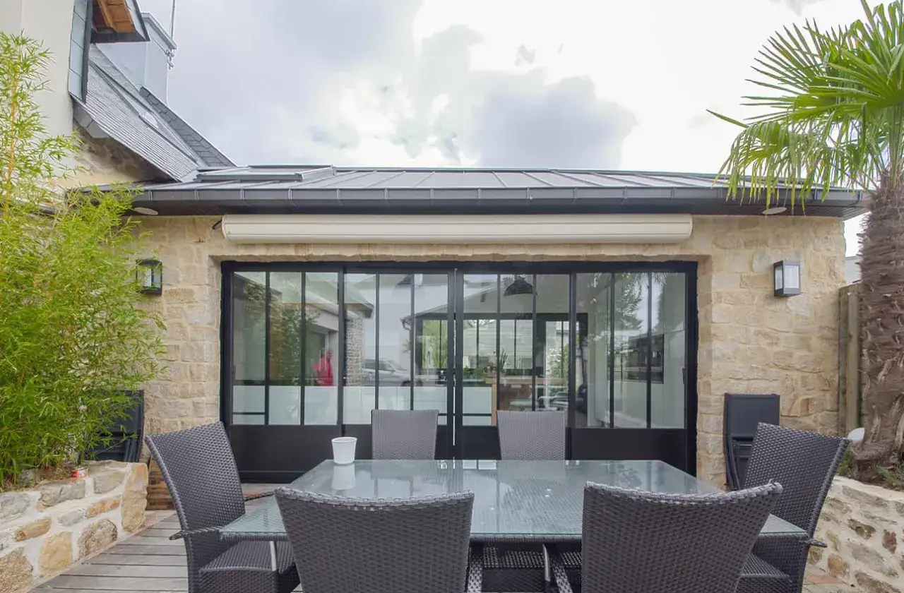 Une verri&egrave;re d'extension de maison moderne avec une table et des chaises en rotin sur une terrasse en bois, entour&eacute;e de v&eacute;g&eacute;tation.