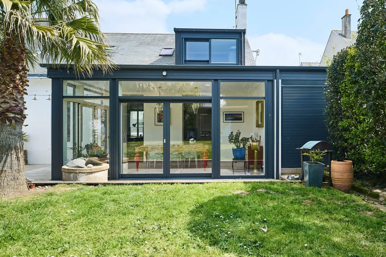 Une extension longère moderne avec de grandes baies vitrées, une palme et un jardin verdoyant.