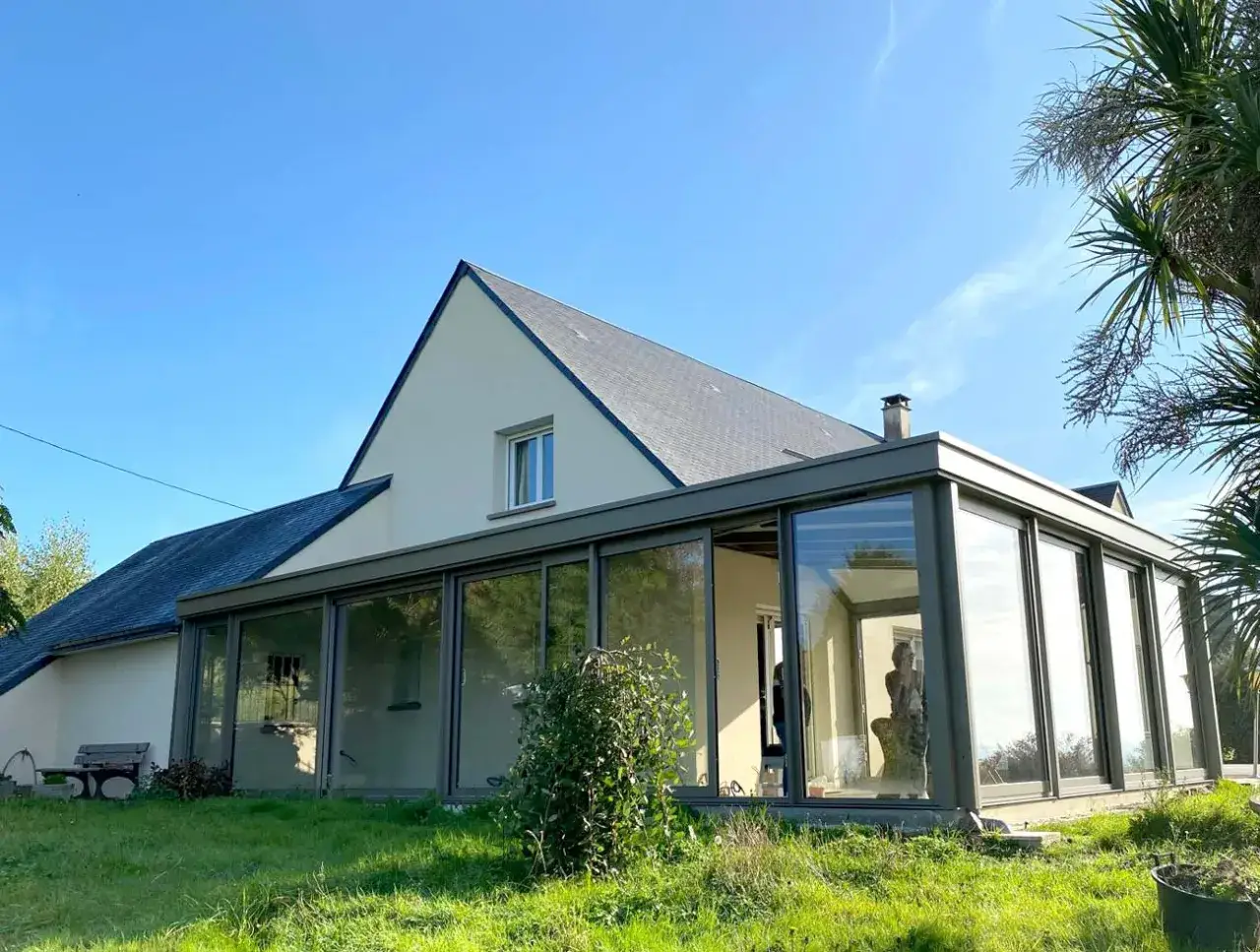 Une maison avec une véranda moderne, dont la toiture est en zinc, s'intègre parfaitement au paysage verdoyant sous un ciel bleu.