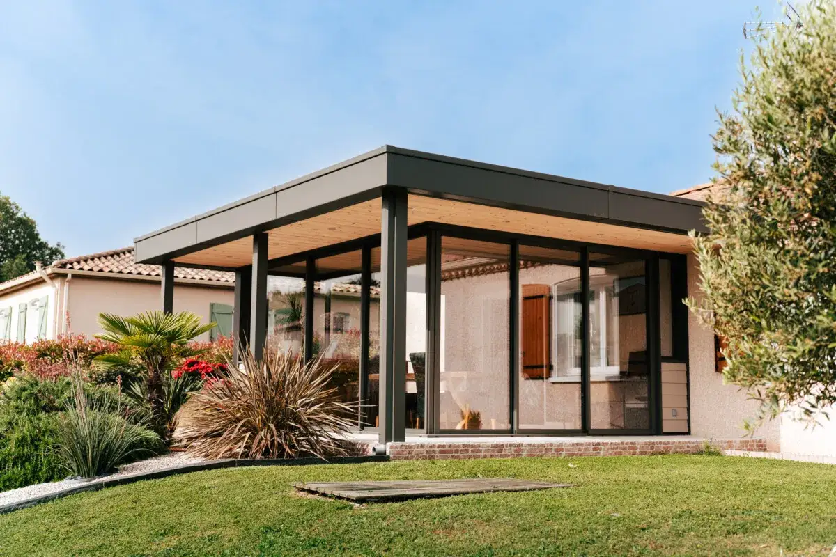 Moderne petite extension de maison avec grandes baies vitrées, toiture en bois et jardin verdoyant.