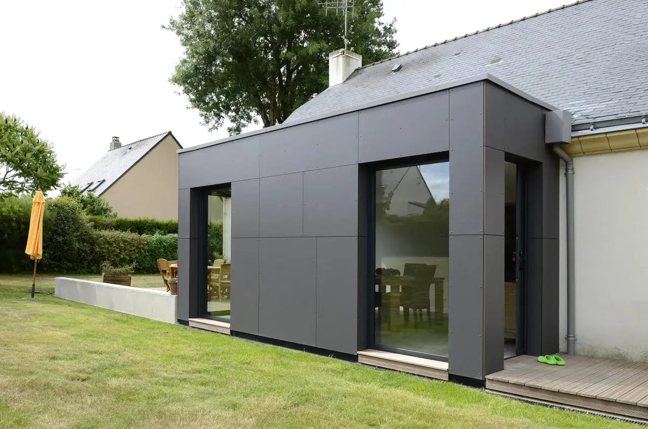 Extension de maison moderne avec fa&ccedil;ade en bac acier et toit plat. Un jardin verdoyant entoure la structure.