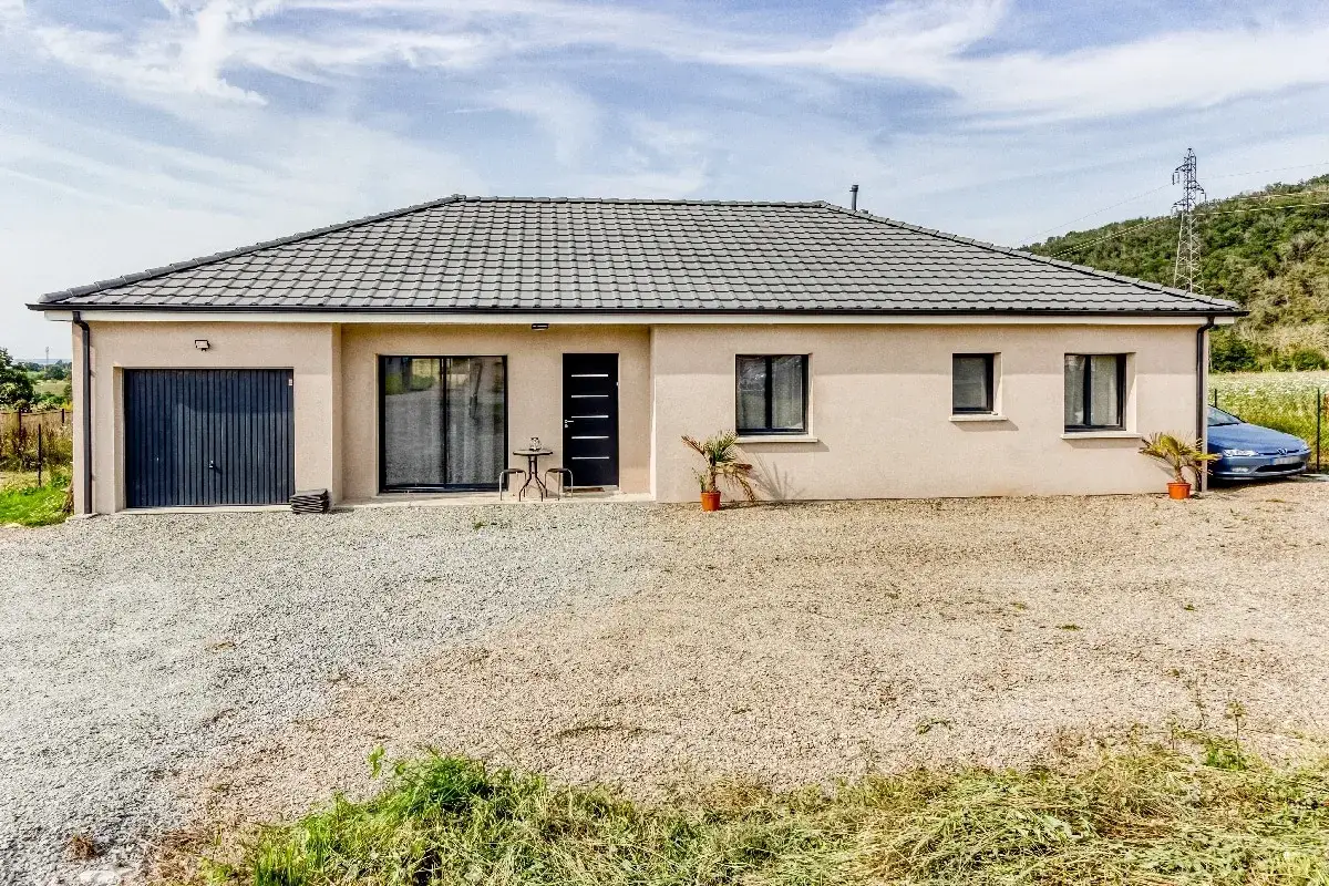 Maison moderne avec garage et un toit 4 pans, devant un paysage verdoyant sous un ciel bleu.