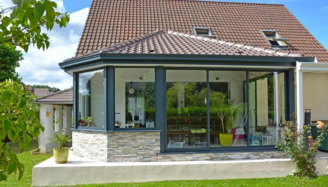 Belle veranda avec toiture tuile, offrant une vue sur un jardin verdoyant. Les grandes baies vitr&eacute;es refl&egrave;tent la nature environnante.