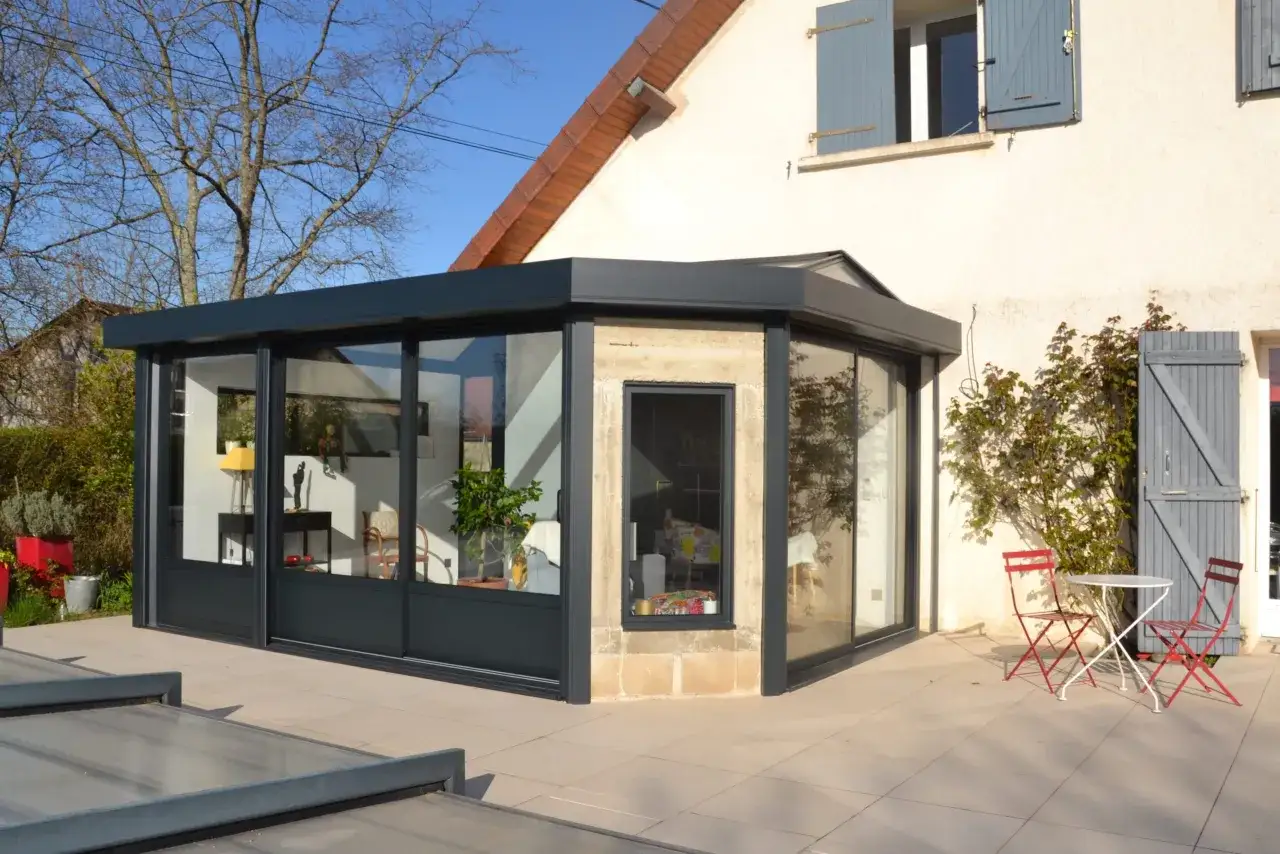 Une v&eacute;randa moderne avec de grandes baies vitr&eacute;es, un petit salon de jardin et des volets bleus. Une extension qui pourrait influencer la taxe fonci&egrave;re.