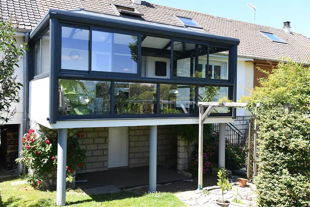 Une &eacute;l&eacute;gante veranda sous balcon, avec une chaise verte visible &agrave; travers les vitres, s'int&egrave;gre parfaitement &agrave; la maison.