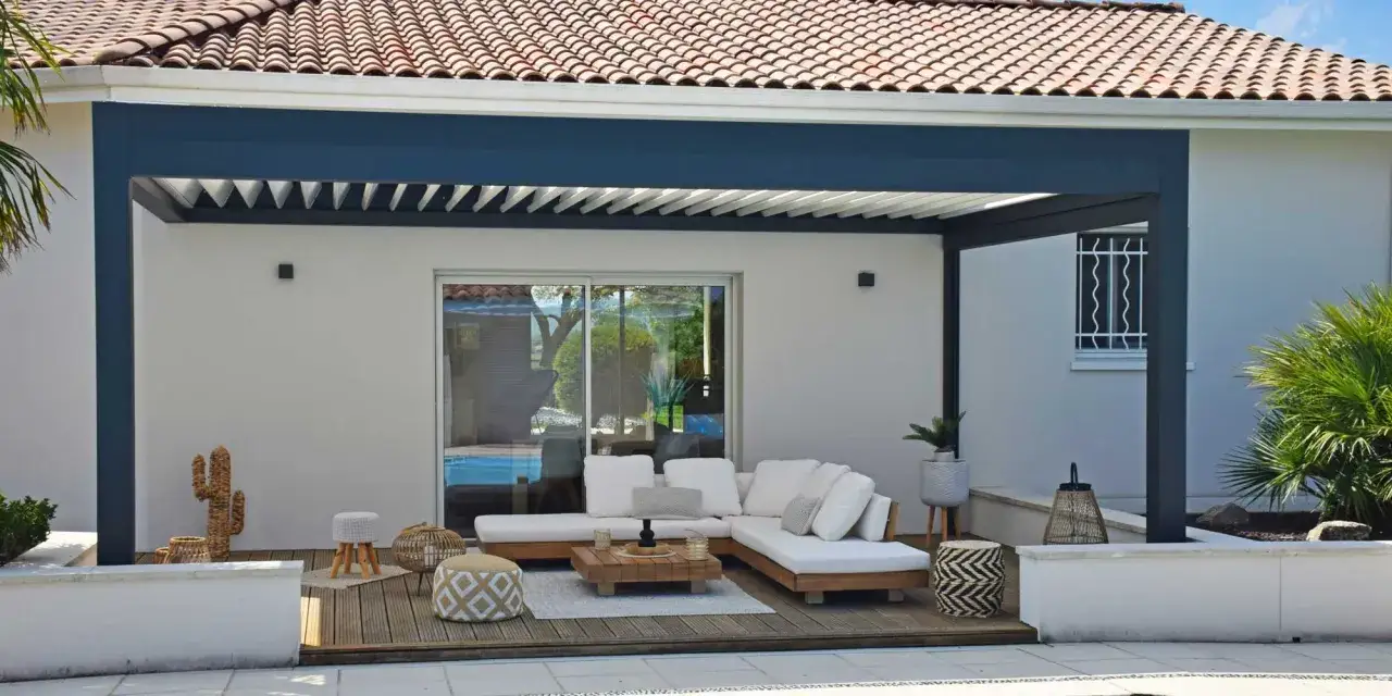 Salon ext&eacute;rieur moderne sous une pergola &agrave; lames orientables. Canap&eacute; blanc, table basse en bois, poufs et cactus d&eacute;coratif cr&eacute;ent une ambiance cosy. La dimension pergola s'int&egrave;gre parfaitement.