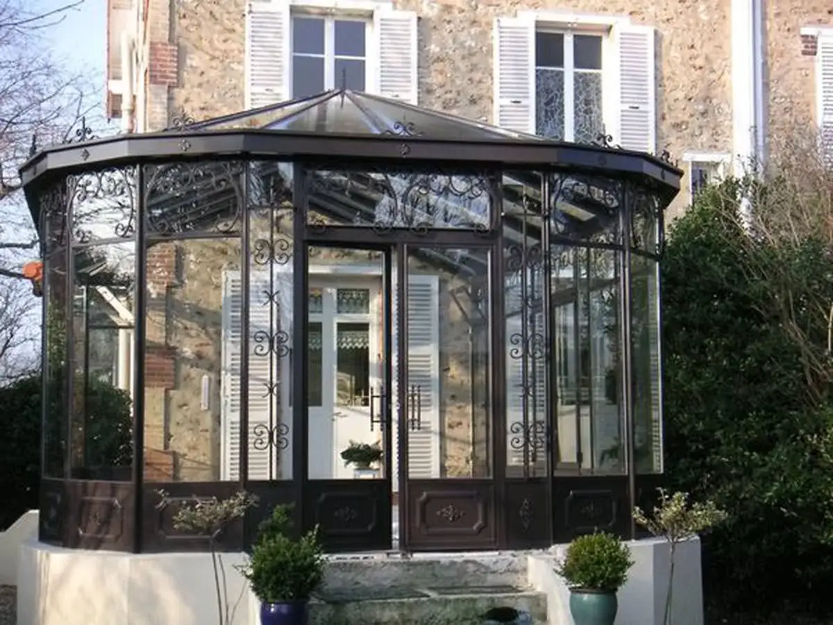 Belle v&eacute;randa couloir en fer forg&eacute; et verre, s'ouvrant sur une maison en pierre. Des volets blancs et des arbustes compl&egrave;tent le d&eacute;cor.