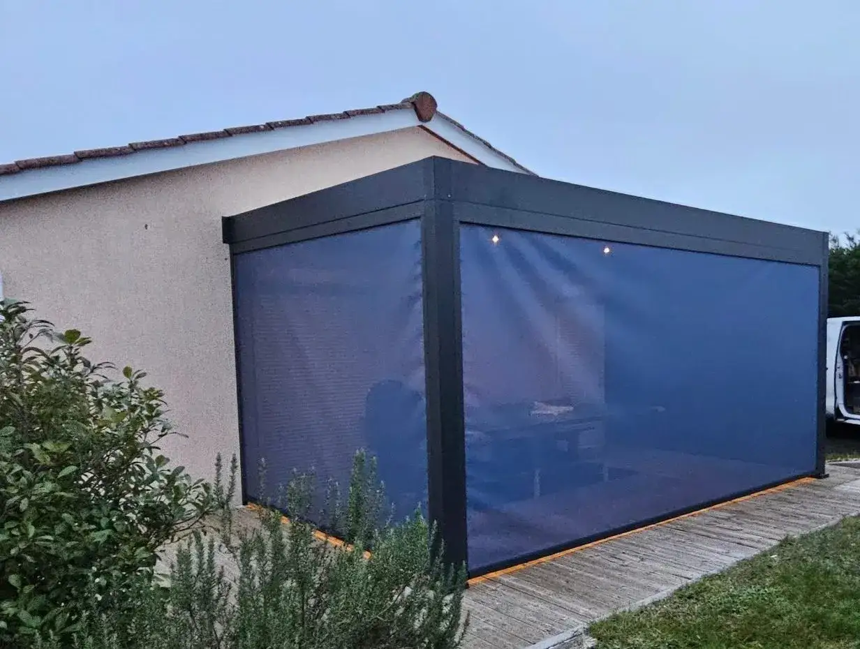 Pergola moderne de moins de 5m2, avec rideaux bleus occultants, installée contre une maison. Un espace cosy pour profiter de l'extérieur.
