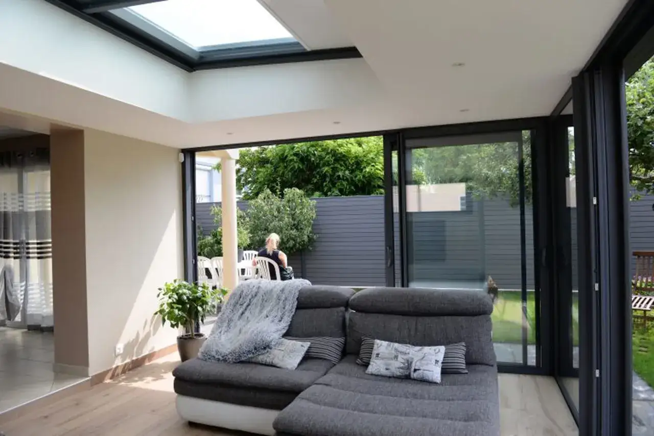 Salon lumineux avec canapé gris, une extension de maison moderne avec toit vitré et grandes baies vitrées donnant sur un jardin verdoyant.