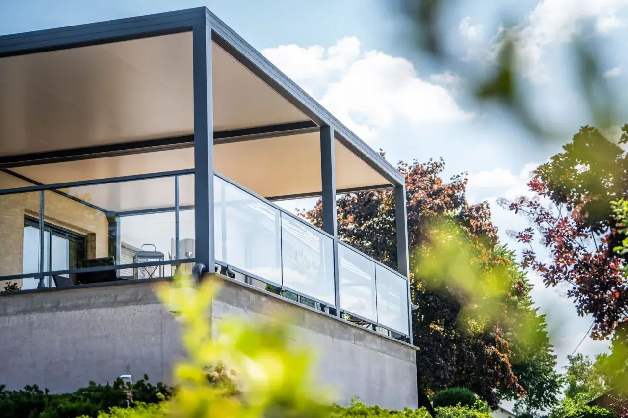 Un &eacute;l&eacute;gant petit veranda balcon avec garde-corps en verre et toit moderne, entour&eacute; d'arbres feuillus.