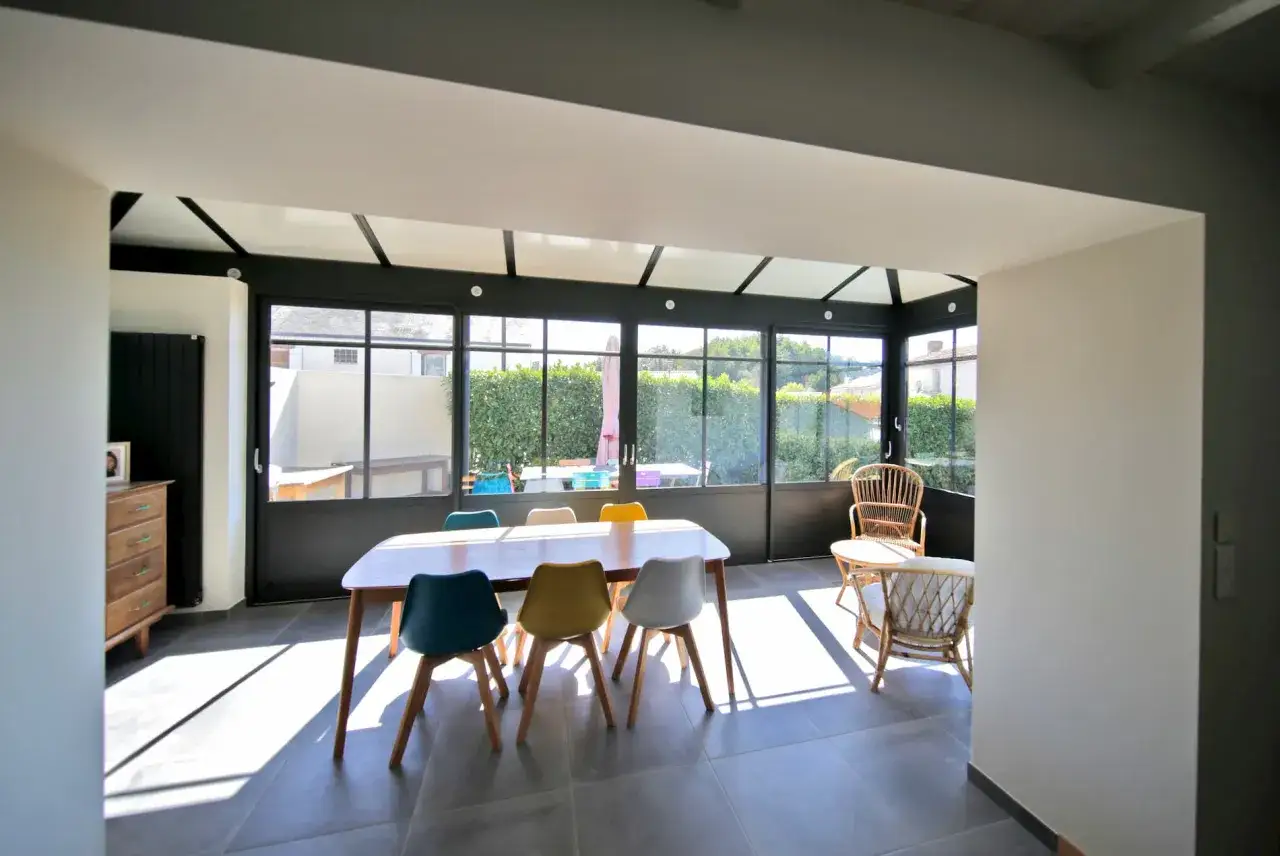 Salle &agrave; manger lumineuse dans une v&eacute;randa chauff&eacute;e, avec une table en bois et des chaises color&eacute;es, offrant une belle surface habitable.