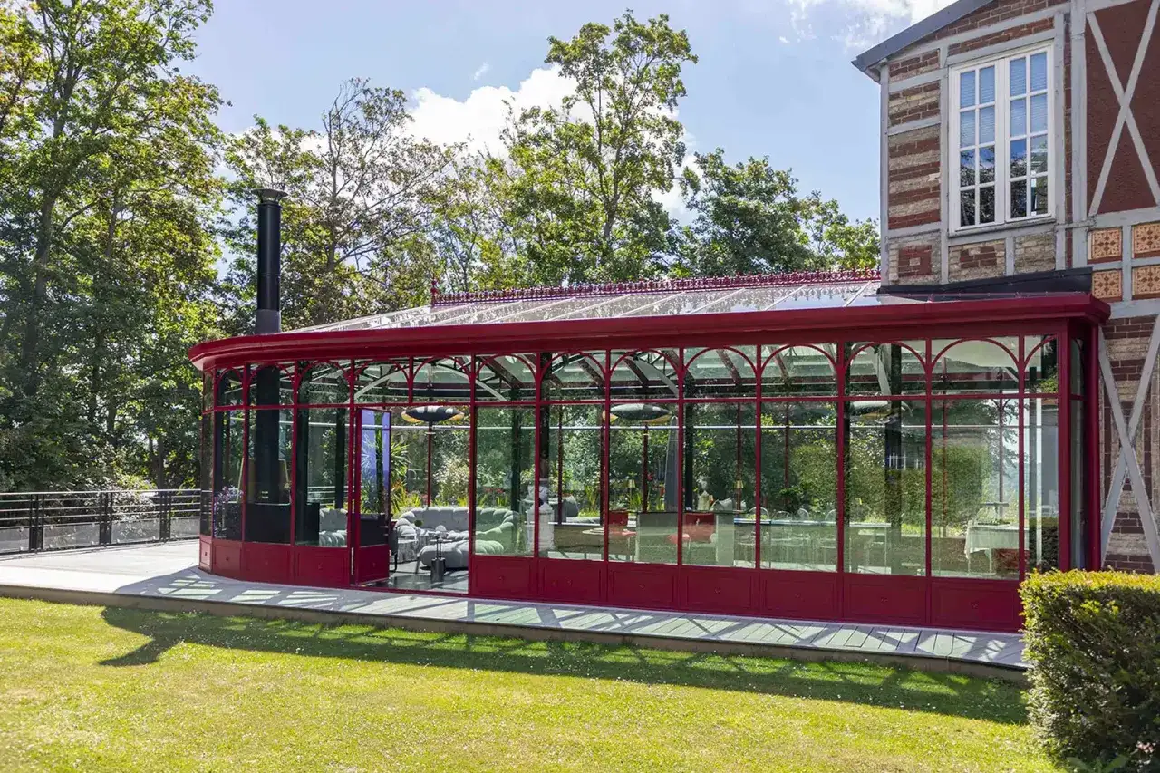 Magnifique jardin d'hiver ancien avec structure rouge, baigné de soleil. Un espace lumineux invitant à la détente, entouré de verdure.