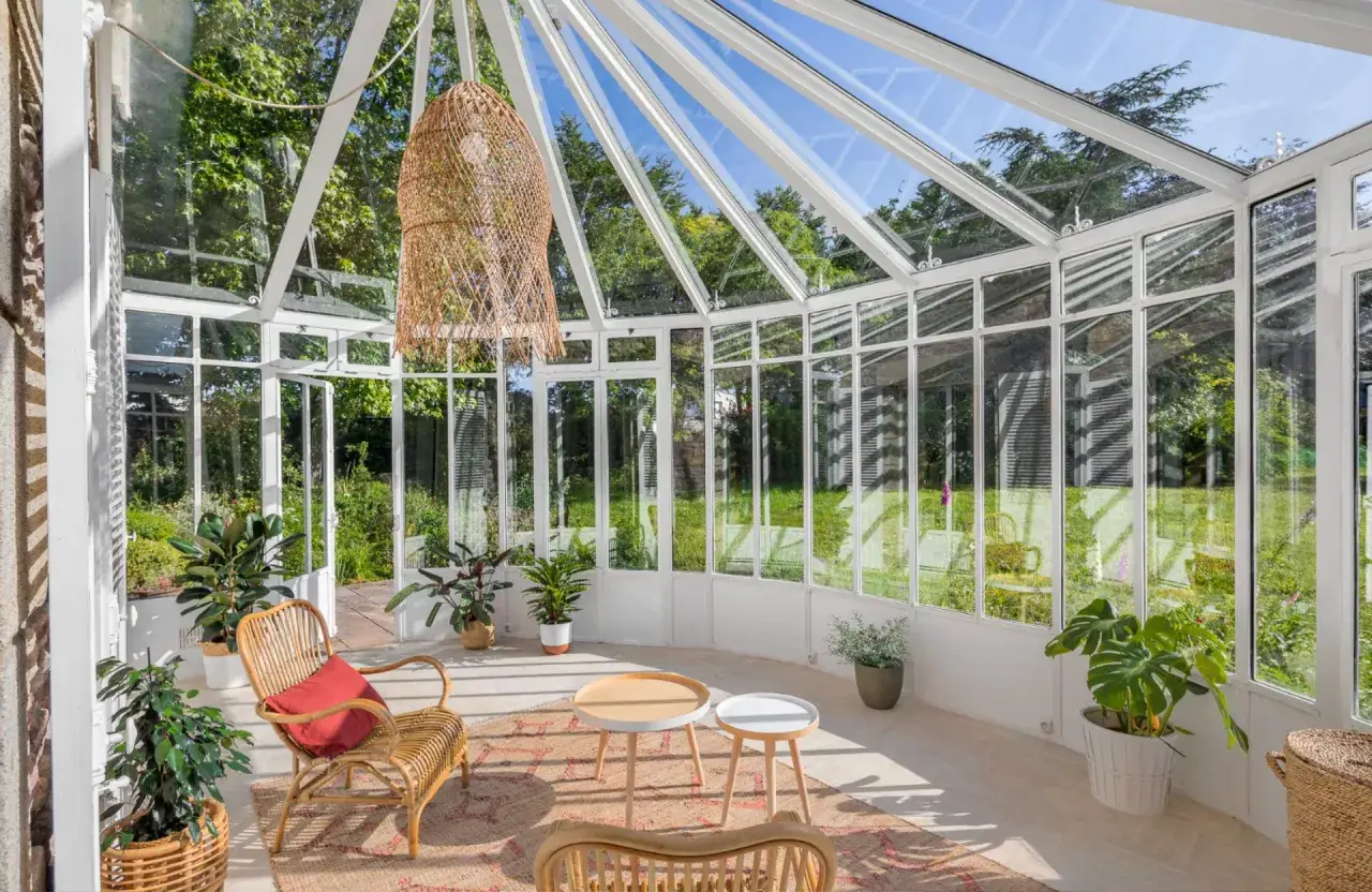 Une véranda en l. lumineuse avec mobilier en rotin, tables basses, plantes vertes et tapis, offrant une vue sur un jardin verdoyant.