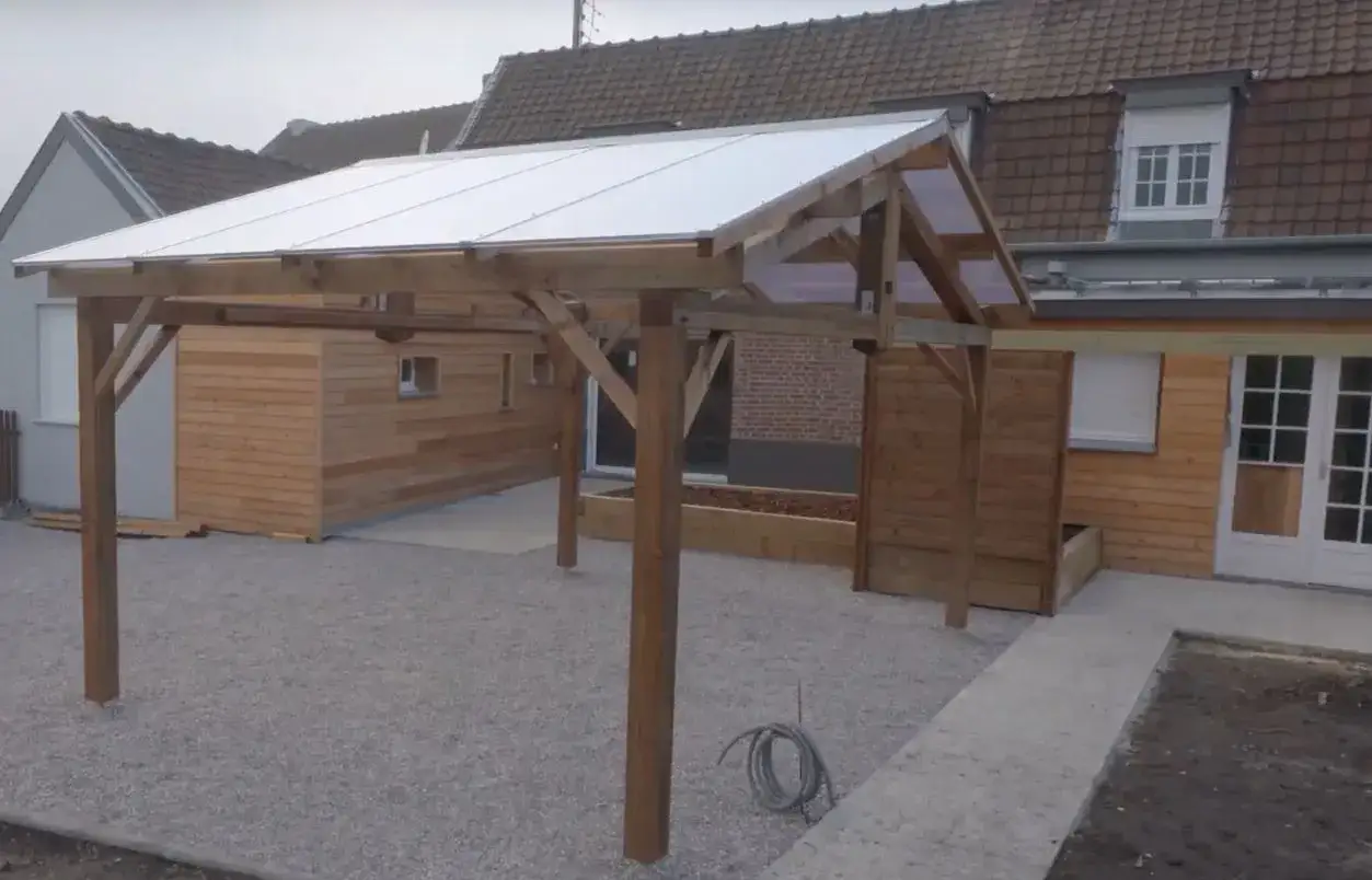 Un carport en bois avec un toit en pente translucide prot&egrave;ge une all&eacute;e en gravier devant une maison en bois.